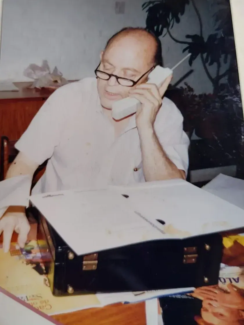 Pedro ao telefone, de camisa branca, com uma pasta aberta sobre a mesa do consultório