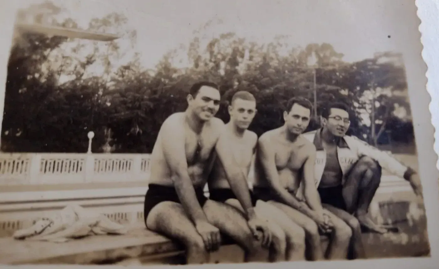 Fotografia antiga de quatro rapazes de sunga sentados na borda de uma piscina, com trampolim e árvores ao fundo