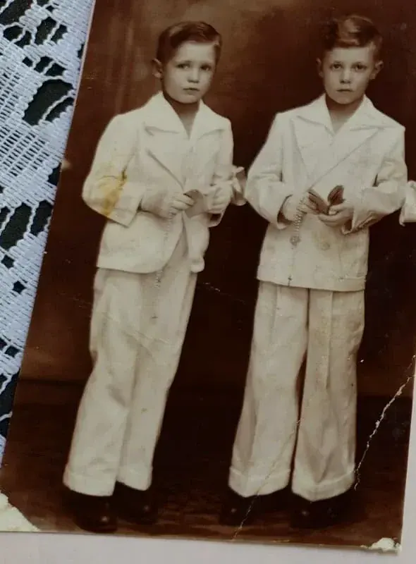 Fotografia sépia de dois meninos de terno branco segurando terços e livrinhos, em traje de primeira comunhão