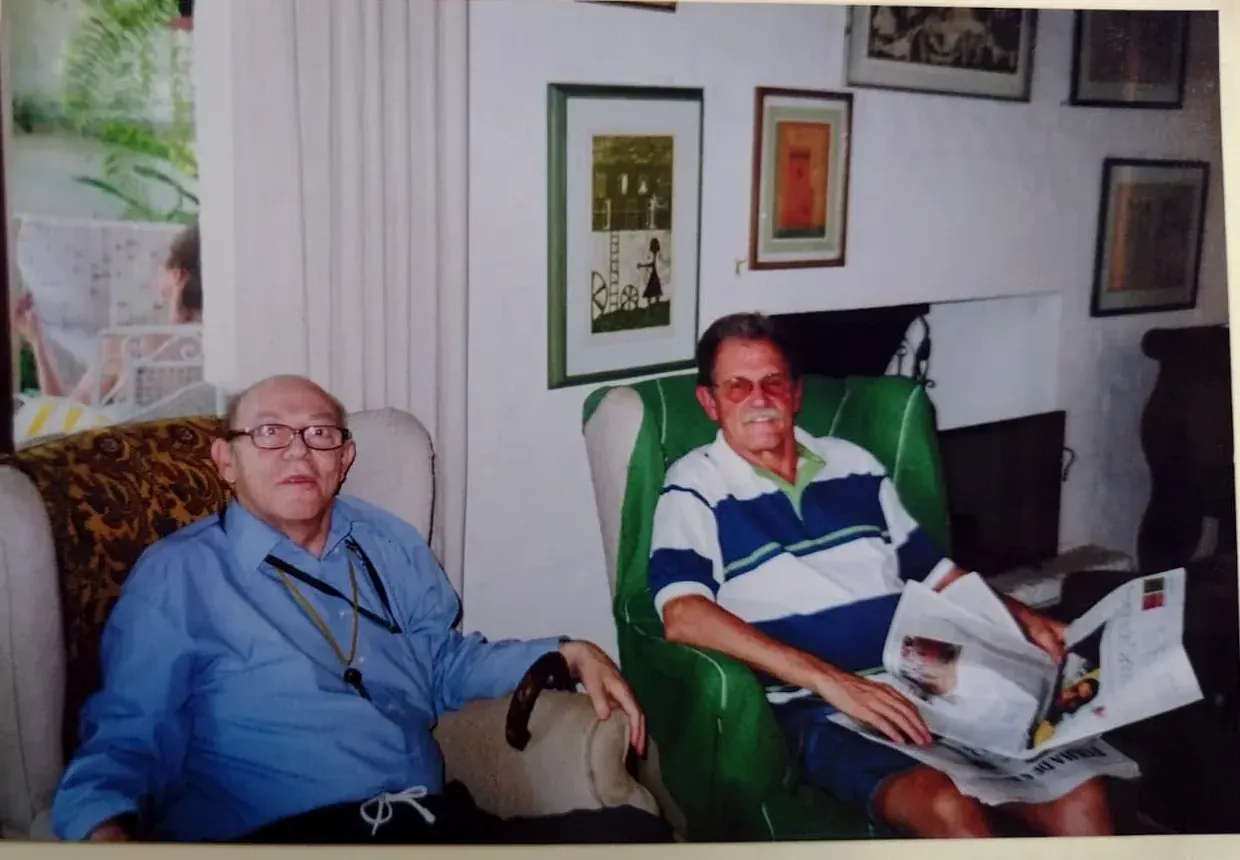 Pedro de camisa azul sentado em uma poltrona ao lado de um amigo que lê o jornal em uma poltrona verde