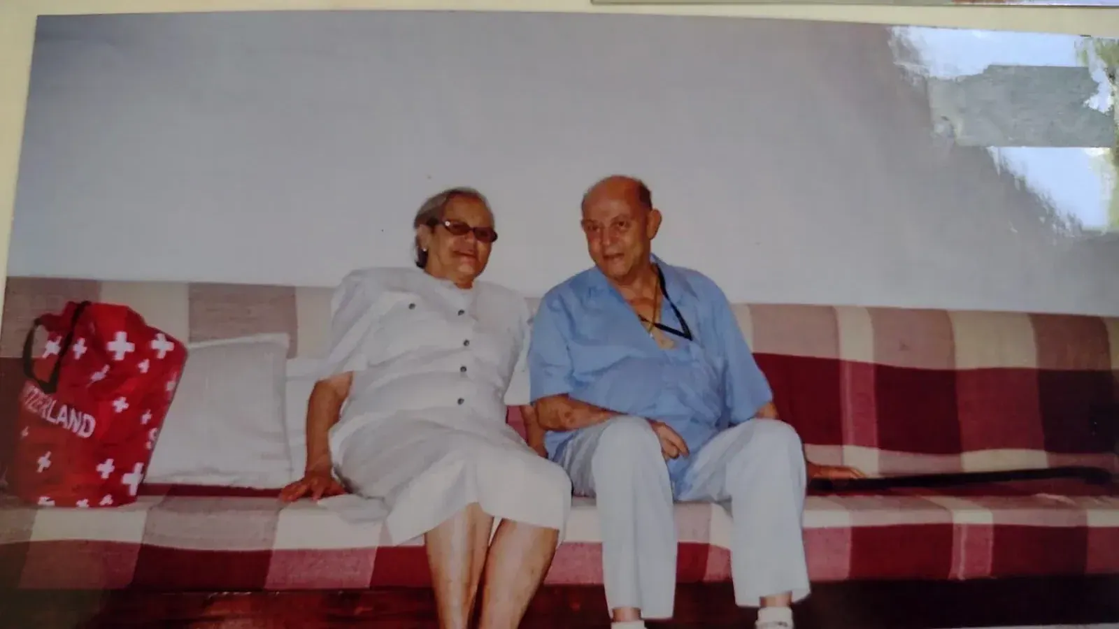Pedro de camisa azul sentado em um sofá xadrez ao lado de uma senhora de branco que sorri