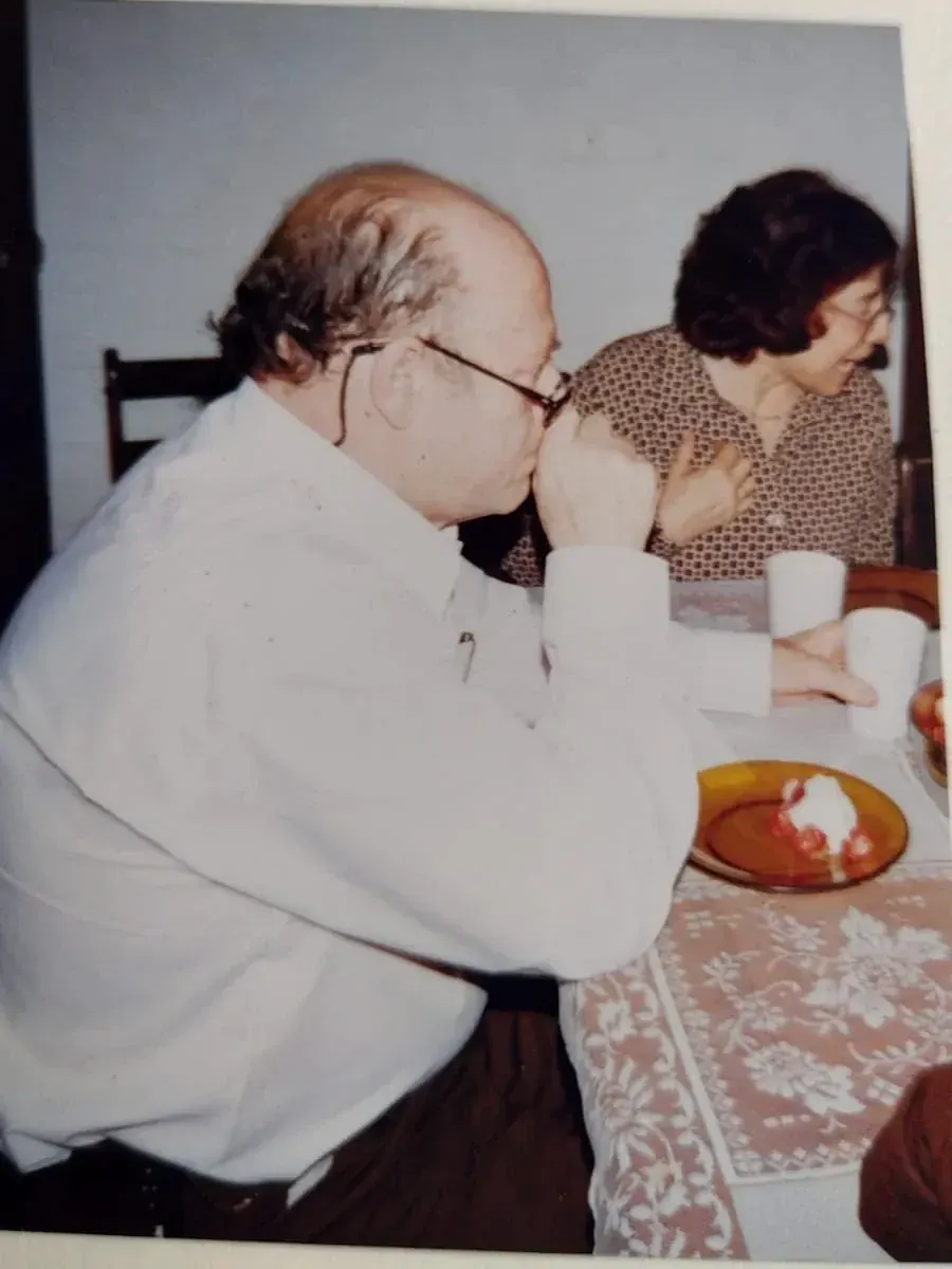 Pedro de perfil, de camisa branca, à mesa com um prato de sobremesa e uma mulher ao lado