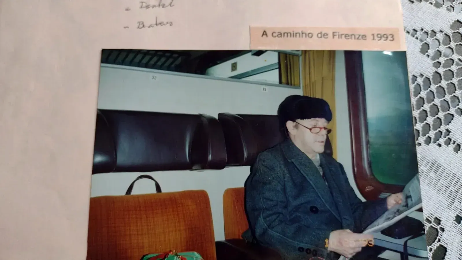 Pedro de boina, casaco cinza e óculos vermelhos lendo um jornal em um trem, com a legenda "A caminho de Firenze 1993"
