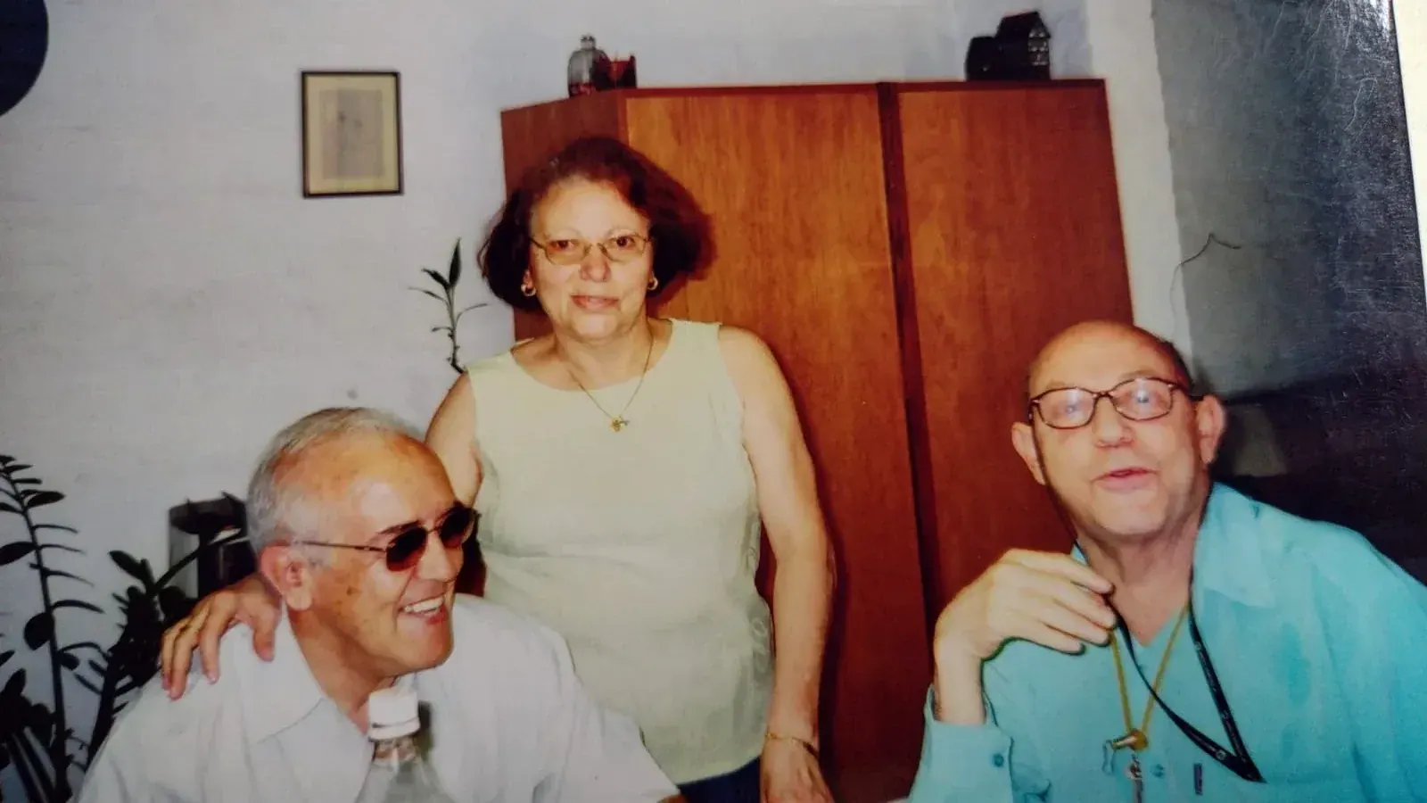 Pedro de camisa verde-água ao lado de um homem sorridente de óculos escuros e uma mulher em pé entre os dois