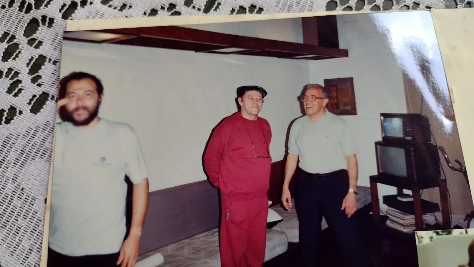 Pedro de agasalho vermelho e boné em pé entre dois homens em uma sala com televisores