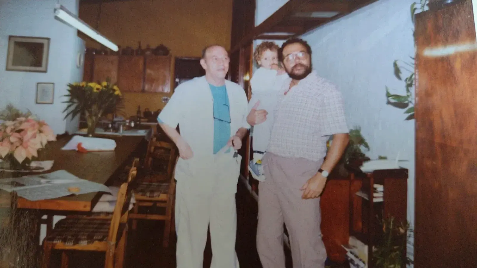 Pedro de jaleco branco em uma cozinha ao lado de um homem de barba que segura uma criança pequena