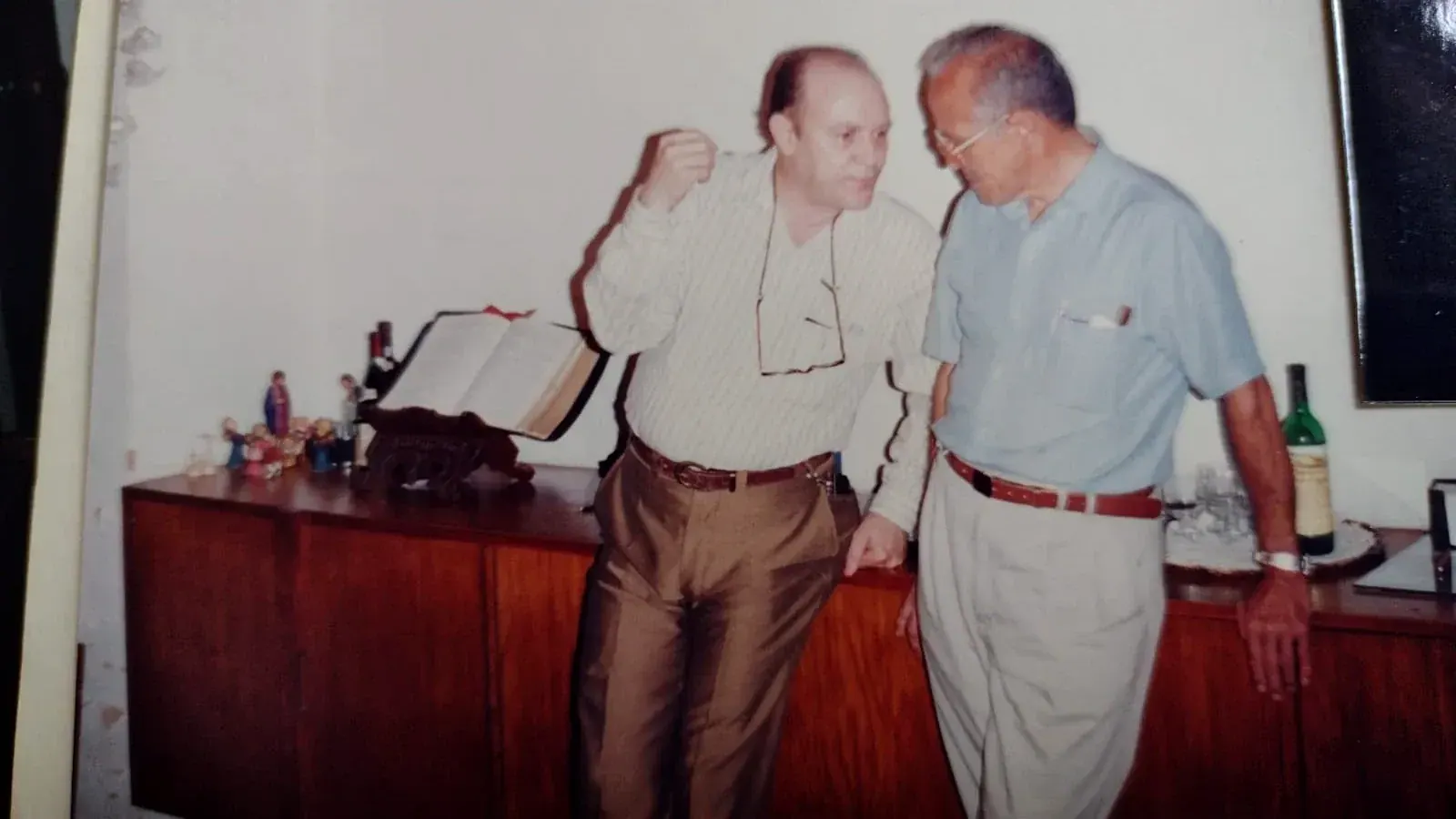 Pedro de camisa listrada conversando com um homem de camisa azul junto a um aparador com um livro aberto e figuras de presépio