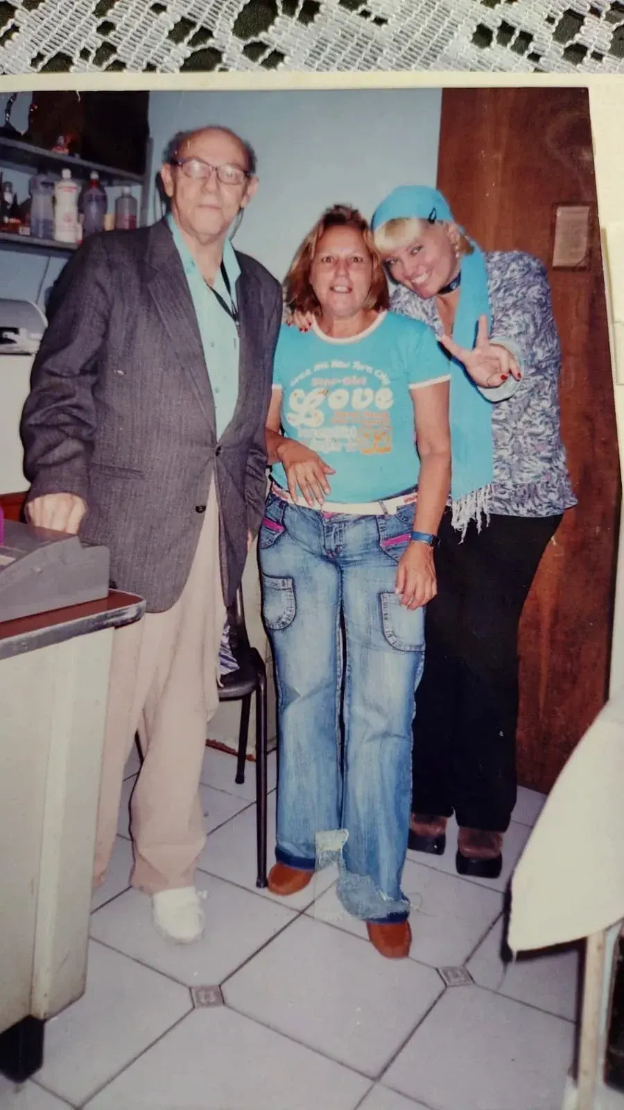 Pedro de blazer cinza sobre camisa azul, sorrindo com duas mulheres em pé em uma sala