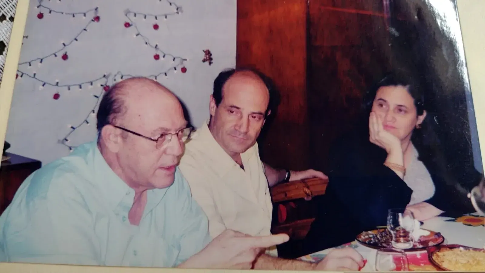 Pedro de camisa verde-clara à mesa de Natal, com um homem e uma mulher ao lado e luzes em forma de árvore na parede