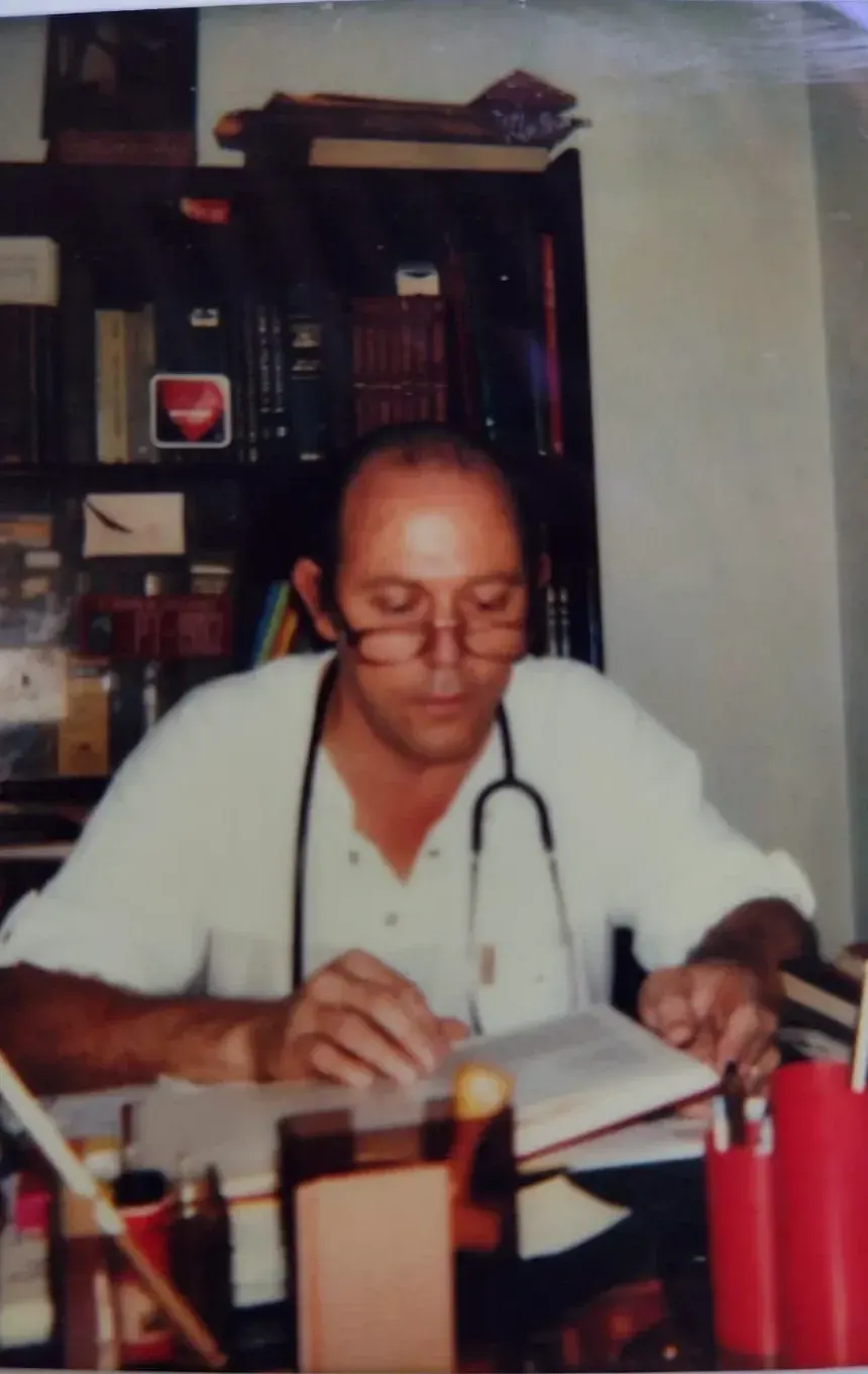 Pedro Moretti Guedes de jaleco branco, com estetoscópio no pescoço, lendo um livro em sua mesa diante de estantes cheias de livros