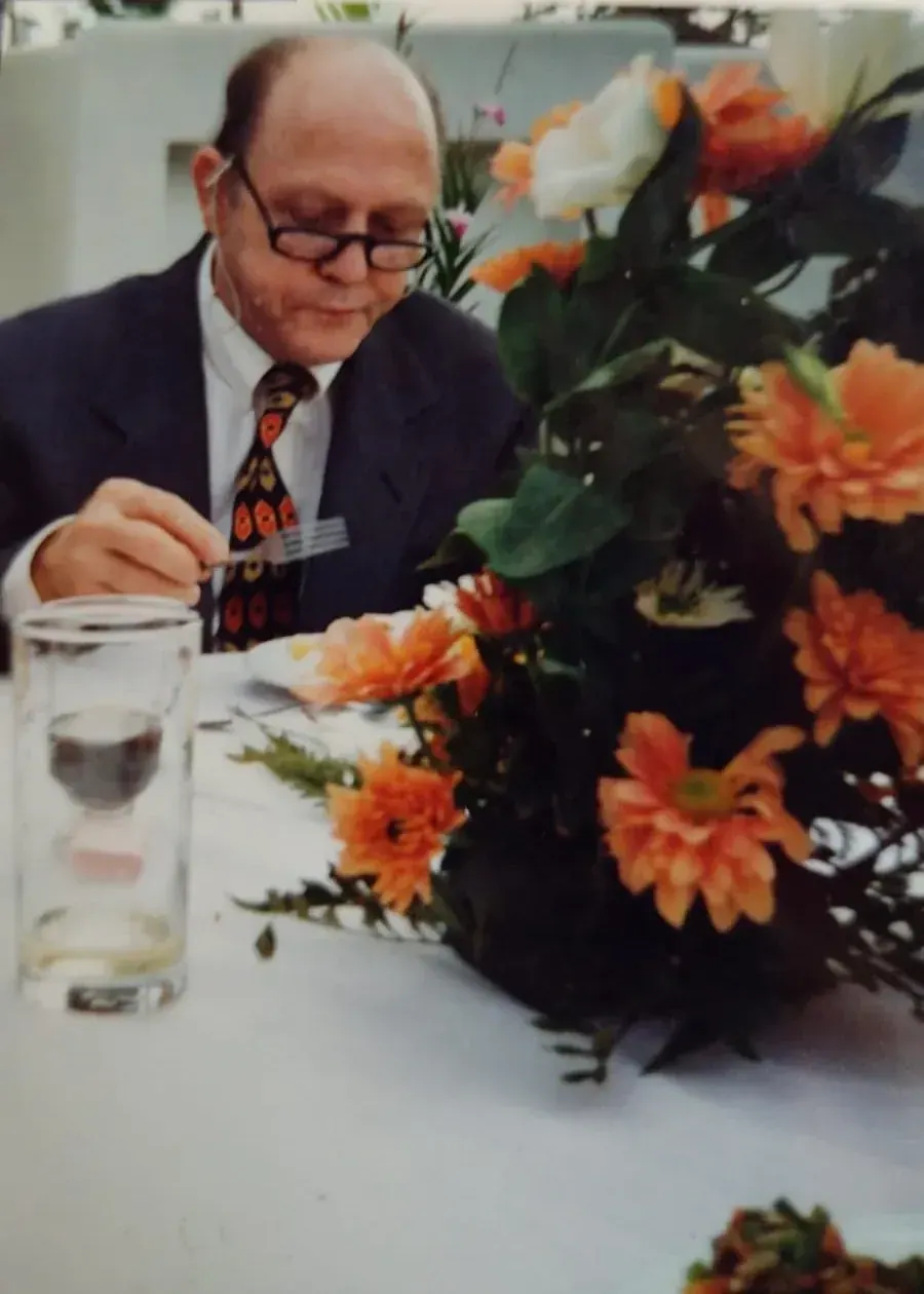 Pedro de terno escuro e gravata estampada em um almoço formal, com um arranjo de flores laranja à frente