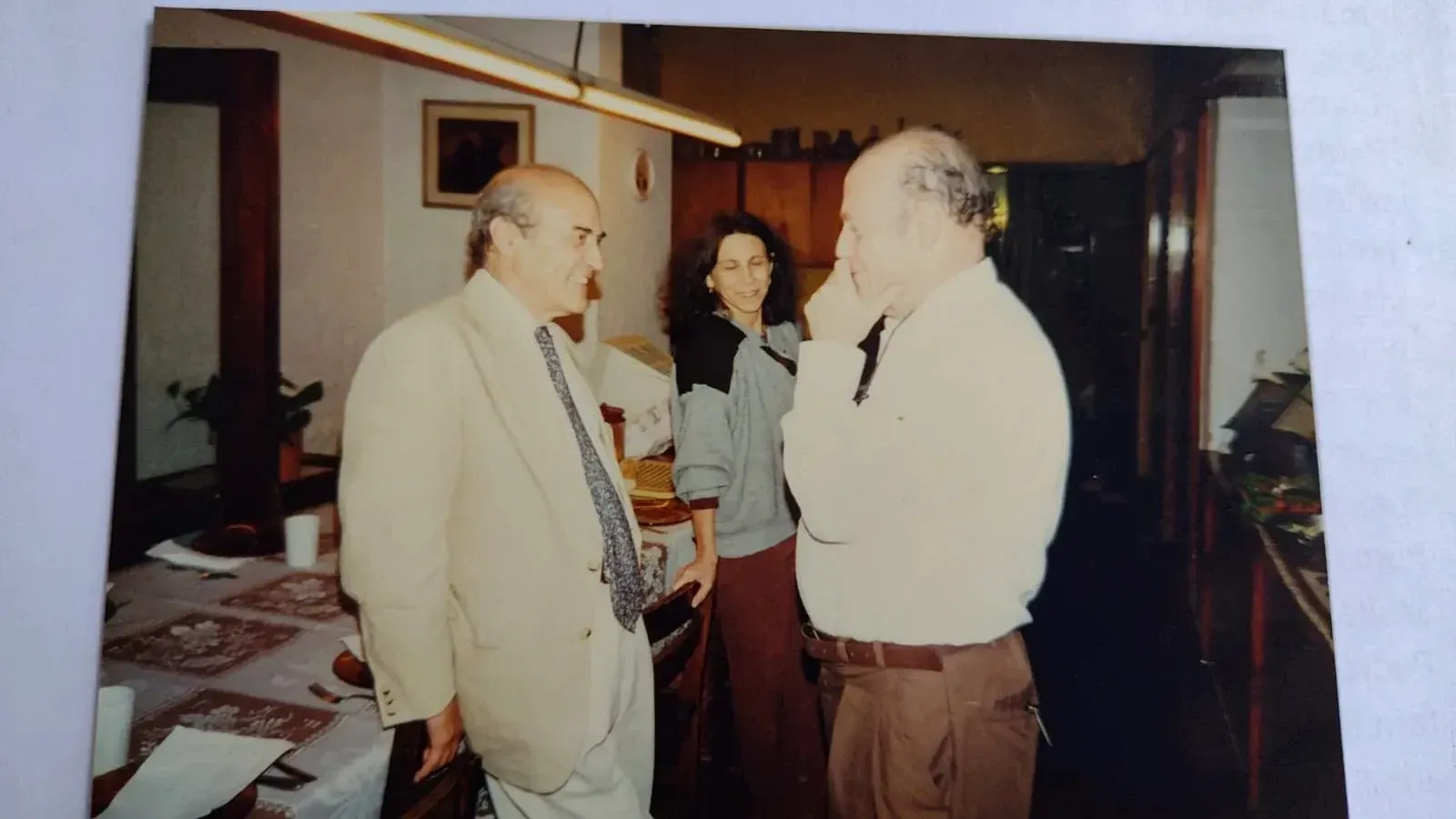 Pedro de suéter claro conversando com um homem de terno bege em uma sala de jantar, com uma mulher sorrindo ao fundo